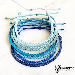 May include: A set of eight braided bracelets in shades of blue, from light blue to dark blue. The bracelets are stacked on top of each other, creating a gradient effect. The bracelets are made of a thin, braided cord.