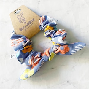 Può includere: Un elastico per capelli blu e bianco con fiori gialli, arancioni e rossi. L'elastico è legato a fiocco e ha un'etichetta che dice "SummerStyle Bracelets".