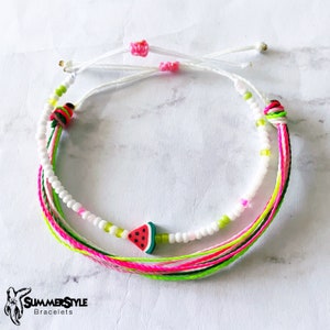 May include: A white and green friendship bracelet with a watermelon charm. The bracelet is made of multiple strands of string and beads.