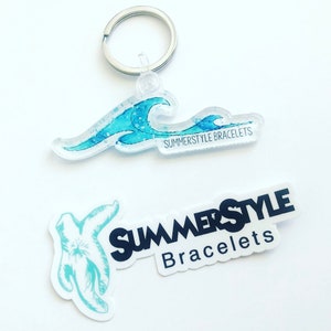 以下が含まれることがあります： 青い波のデザインと「SUMMERSTYLE BRACELETS」というテキストが書かれた透明なアクリルキーホルダー。別途、「SUMMERSTYLE Bracelets」というテキストが書かれたステッカーも表示されています。