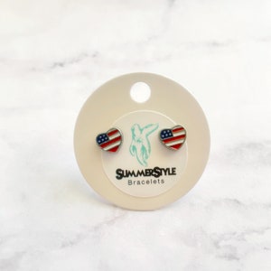 Könnte beinhalten: Ein Paar silberne herzförmige Ohrstecker mit amerikanischem Flaggenmuster. Die Ohrringe werden auf einer weißen Karte mit dem Text "SummerStyle Bracelets" gezeigt.