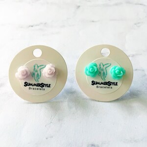 May include: Two pairs of flower stud earrings on white cards. The earrings are made of plastic and are in the shape of a rose. One pair is light pink and the other pair is turquoise. The cards have the text "SummerStyle Bracelets" printed on them.