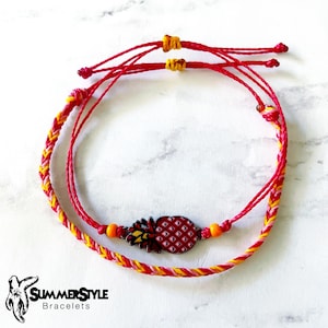 Puede incluir: Dos pulseras de amistad rojas y amarillas con un dije de piña. El dije es rojo, amarillo y blanco con un contorno negro. Las pulseras están atadas juntas con un nudo.