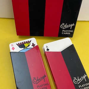 May include: A set of playing cards in a red, black, and white box. The box has the text "Obergs PLAYING CARDS". Two card decks are visible, one with a King card and the other with an Ace card. The box is striped.