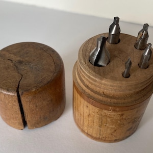 May include: A set of five small metal drill bits in a wooden case. The case is made of light brown wood and has a lid that fits snugly over the drill bits. The drill bits are arranged in a circle around the center of the case.