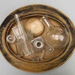May include: An assortment of clear glass laboratory equipment and bottles, including a 200 ml Pyrex flask, arranged inside a distressed oval wooden tray with a brown interior and black rim. The collection includes various shapes and sizes.