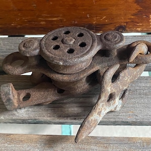 May include: A heavy, rusted iron pulley block featuring a circular wheel with six small holes, a metal frame, and an attached hook. The surface of the object shows significant oxidation and a dark brown, weathered texture throughout the metal structure.