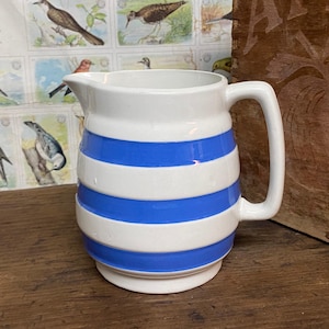 May include: White ceramic pitcher with blue horizontal stripes. The pitcher has a curved handle and a spout for pouring. The background features a vintage bird illustration.