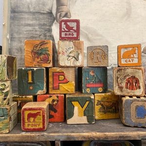 May include: A collection of vintage wooden alphabet blocks featuring colorful illustrations of animals, objects, and letters. The blocks display images of a bird, rabbit, cat, pony, cow, and a child with a wagon, alongside letters like P, Y, and C.