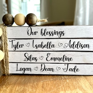 May include: White wooden sign with the text "Our blessings" on the top line, followed by three lines of names with hearts between each name: Tyler, Isabella, Addison, Silas, Emmeline, Logan, Dean, Jade. The sign is decorated with a brown and beige beaded garland.