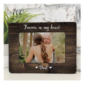May include: A rectangular dark brown wooden picture frame with a distressed finish. The top of the frame features the white text "Forever in my heart" and the bottom features the white text "Dad" flanked by two small white heart icons.
