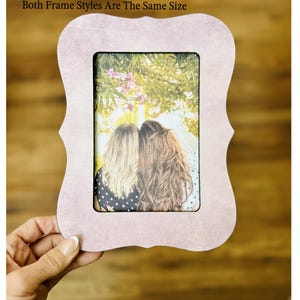 Op de afbeelding: Een lichtroze, decoratieve fotolijst met een unieke, gebogen vorm. De lijst bevat een foto van twee mensen met lang haar. Afmetingen lijst: 21,6 cm x 16,5 cm, foto-opening: 10,2 cm x 15,2 cm. De tekst "Words Worth Framing Co" is zichtbaar.