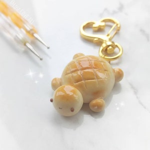 May include: A small, golden brown turtle keychain made of polymer clay. The turtle has a detailed shell with a grid pattern and a cute, smiling face. The keychain is attached to a gold heart-shaped clasp.