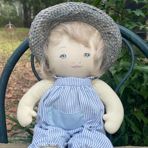 May include: A handmade doll with a straw hat, blonde hair, and blue eyes. The doll wears blue and white striped overalls and matching shoes. The doll is sitting on a wooden surface.