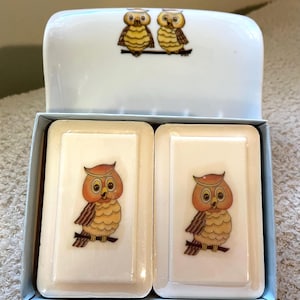 May include: A vintage soap dish set featuring a white lid with two owl illustrations and two rectangular soap bars. Each bar has a single owl illustration in orange, brown, and yellow.
