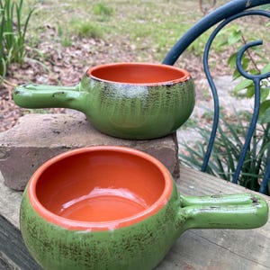 Puede incluir: Dos cuencos de sopa de cerámica con asas. El exterior es verde texturizado, mientras que el interior es de color naranja brillante. Los cuencos son redondos con un solo asa en el lateral. Están sobre una superficie de madera.