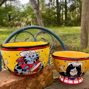 May include: Two yellow ceramic bowls with cat chef designs. The larger bowl features a blue cat chef with a red apron, while the smaller bowl has a black and white cat chef. Both bowls have a red rim and are decorated with blue polka dots.
