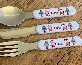 May include: A set of three Christmas-themed children's cutlery with a white handle featuring a Santa Claus, reindeer, and Christmas tree design. The handle is personalized with the name "Charlie".
