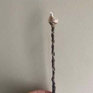 May include: A handcrafted wand with a natural wood shaft wrapped with twine. The top features a light-colored, bulbous element, possibly a crystal or stone, secured with twine. The wand is approximately 10 inches long.