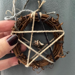 May include: A small, circular wreath made of woven brown twigs, with a five-pointed star design created by beige and navy blue twine. A small seashell is nestled in the center. The wreath is adorned with gray twine for hanging.