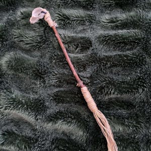 May include: A handcrafted wand with a clear crystal at the top. The wand's handle is a natural brown stick, wrapped with beige twine. The bottom of the wand has a fringe of beige twine. The wand is set against a dark green, textured background.