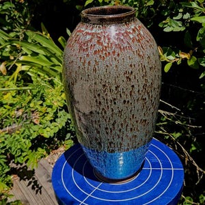May include: A ceramic vase with a tapered neck and a rounded body. The upper portion features a mottled brown and grey glaze with vertical drip patterns, while the lower section transitions into a solid, vibrant blue glaze at the base.
