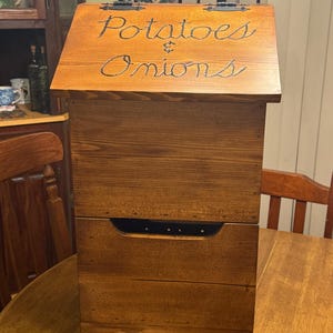 May include: Wooden potato and onion storage bin with a hinged lid. The lid is inscribed with the words "Potatoes & Onions" in a cursive font. The bin has a drawer with a black handle and is made of stained wood.