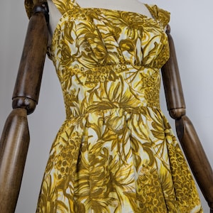 May include: A vintage-style dress with a yellow and white floral pattern. The dress features a square neckline, wide shoulder straps, and a fitted bodice that flares into a full skirt. The fabric has a textured appearance, and the dress is displayed on a mannequin.
