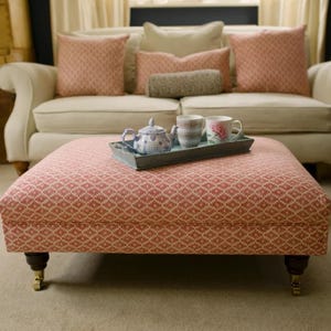 May include: A red and white patterned ottoman with a tray holding a teapot and cups. The ottoman has wooden legs with brass castors. Cushions in pink and beige tones are arranged on a cream sofa in the background. The ottoman is approximately 90 cm long.