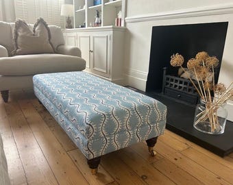 May include: A rectangular ottoman upholstered in light blue fabric with a repeating dark grey wave pattern. The piece features four dark wooden legs with brass casters, providing a stable base for this functional piece of furniture.