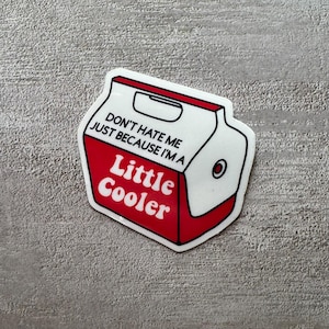 May include: A die-cut sticker shaped like a portable beverage cooler. The cooler is red and white with a handle cutout at the top. It features the black text "DON'T HATE ME JUST BECAUSE I'M A" and the red text "Little Cooler" printed on the front.