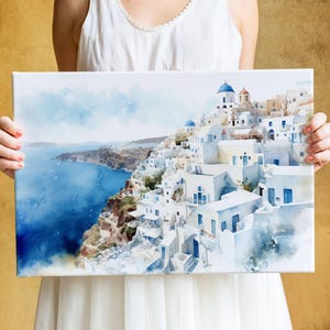 A woman holds a framed painting of a picturesque coastal town with white buildings and blue roofs, overlooking the ocean.
