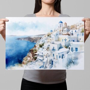A woman holds up a framed print of a picturesque coastal town with white buildings and blue roofs, overlooking the ocean.