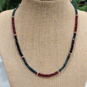 May include: A necklace featuring dark green, dark red, and black beads, interspersed with gold-coloured spacers. The necklace is presented on a tan bust.