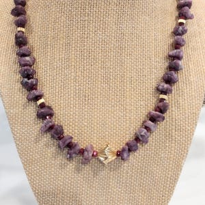 May include: A necklace featuring rough-cut, deep purple gemstones. The necklace has gold-colored spacers and a central gold-colored, geometric pendant. The necklace is displayed on a burlap-covered bust.