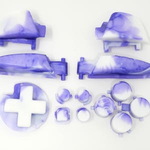 K&ouml;nnte beinhalten: Eine Sammlung von Ersatzteilen f&uuml;r Gamecontroller in einem marmorierten lila und wei&szlig;en Design. Das Set enth&auml;lt zwei Schultertasten, zwei Trigger, ein Steuerkreuz und verschiedene Knopfformen. Die Teile sind auf einer wei&szlig;en Oberfl&auml;che angeordnet.