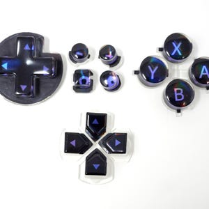 May include: A collection of iridescent black and purple video game controller buttons. The set includes a directional pad, action buttons labeled X, Y, A, and B, and other control buttons. The buttons have a glossy finish and are arranged on a white surface.