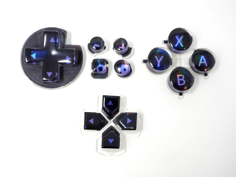 May include: A collection of iridescent black and purple video game controller buttons. The set includes a directional pad, action buttons labeled X, Y, A, and B, and other control buttons. The buttons have a glossy finish and are arranged on a white surface.