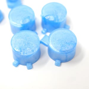 May include: Close-up of several light blue, cylindrical objects with a textured surface and small square feet. The objects are arranged on a white background. The items appear to be small containers or components.