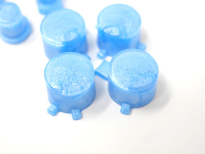 May include: Close-up of several light blue, cylindrical objects with a textured surface and small square feet. The objects are arranged on a white background. The items appear to be small containers or components.