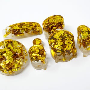 May include: A collection of clear resin objects embedded with gold leaf flakes. The set includes a round piece, a rectangular piece, and several other uniquely shaped pieces. The gold flakes shimmer against the clear resin.