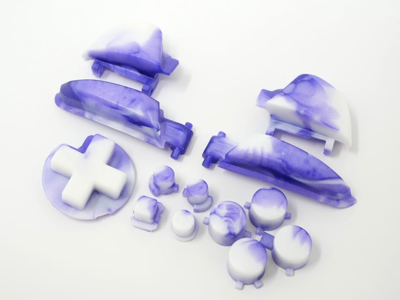 K&ouml;nnte beinhalten: Eine Sammlung von Ersatzteilen f&uuml;r Gamecontroller in einem marmorierten lila und wei&szlig;en Design. Das Set enth&auml;lt ein Steuerkreuz, Tasten und Schultertasten. Die Teile sind zur optischen Aufwertung einer Spielkonsole gedacht.