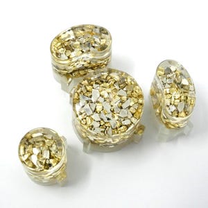 May include: A collection of clear resin jewelry pieces filled with gold and silver-colored fragments. The set includes a ring, a pair of earrings, and two other decorative pieces. The jewelry is displayed on a white surface.