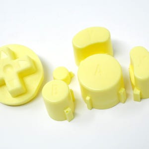May include: A selection of pale yellow video game controller buttons and a directional pad. The buttons include an A button, a B button, a Y button, and a directional pad. The buttons are made of plastic and are on a white background.