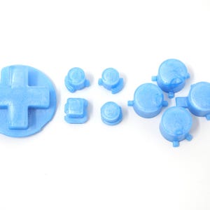 May include: A set of light blue gaming controller replacement buttons, including a D-pad, round buttons, and square buttons. The buttons are made of a solid, opaque material and are arranged on a white background.