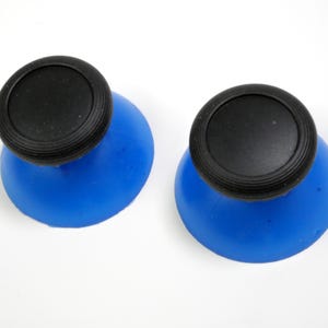 Może przedstawiać: Dwa niebieskie korki do butelek z czarnymi wierzchami. Korki mają zaokrąglony kształt z teksturowanym czarnym wierzchem. Niebieska podstawa jest gładka i zakrzywiona. Korki znajdują się na białym tle.