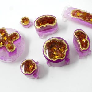 May include: A collection of handmade, resin-cast game controller buttons in various shapes. The buttons are primarily translucent purple with gold-colored accents and a textured, reddish-brown center. The shapes include a cross, circles, and oblongs.