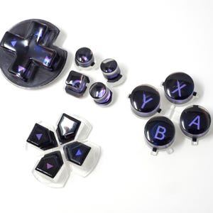 May include: A collection of iridescent purple and black video game controller buttons. Includes a directional pad, various button shapes, and buttons labeled with letters and symbols. The buttons are set against a white background.