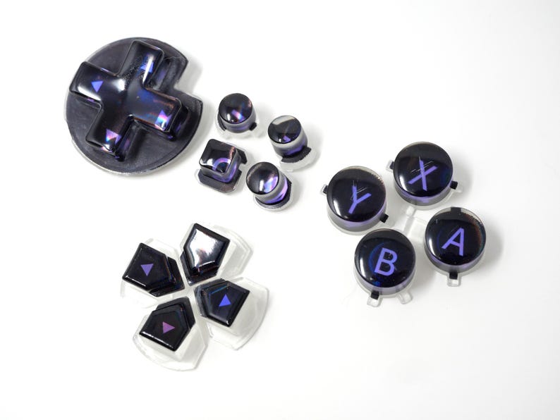 May include: A collection of iridescent purple and black video game controller buttons. Includes a directional pad, various button shapes, and buttons labeled with letters and symbols. The buttons are set against a white background.