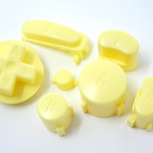 May include: A selection of pale yellow gaming console buttons, including a directional pad, a long rectangular button, and several buttons labelled with letters. The buttons are made of plastic and are arranged on a white surface.